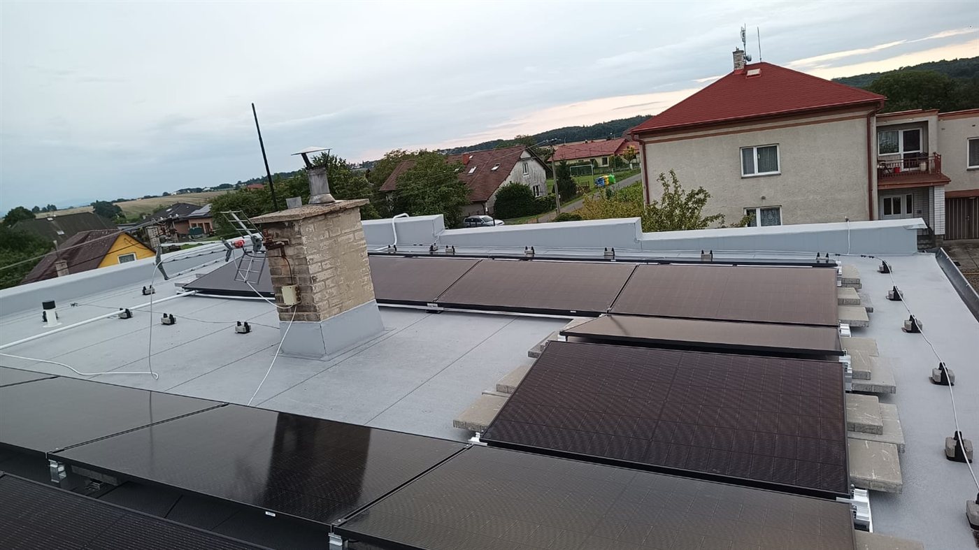 Řady černých panelů na ploché střeše, v pozadí obec